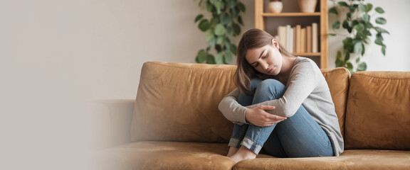 Obraz premium Young woman sits on brown couch, head bowed, knees hugged, conveying sadness or contemplation, ample free space for text