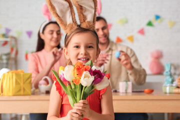 Happy little girl in bunny ears holding tulips and her parents painting Easter eggs at home