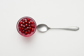 A glass bowl of fresh pomegranate seeds next to a silver spoon on a white background, showcasing vibrant red and shiny metallic colors.