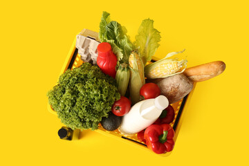 Bottle of oil, yellow shopping basket full of different fresh products and lettuce on yellow background. Top view