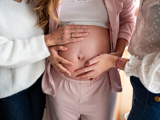 Three women gather to celebrate an upcoming arrival, placing their hands on a visibly pregnant...
