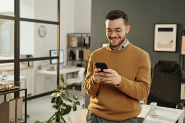 Waist up portrait of smiling young man using smartphone while standing in modern office setting and...
