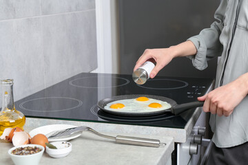 Woman adding salt into frying pan while cooking eggs on cooktop in kitchen, closeup
