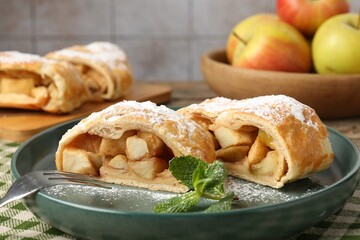 Pieces of tasty apple strudel with powdered sugar, mint and fork on table against grey tiled wall, closeup