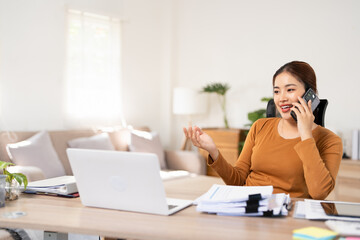 Calling clients on a mobile phone. Professional business woman looking at a laptop screen while talking on a phone call. Young business woman working in home office.