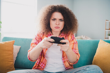 Young woman playing video games while sitting on a couch in a cozy living room, expressing focus...