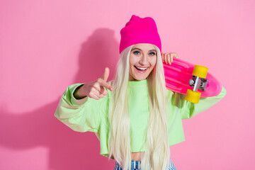 Cheerful young woman posing in stylish casual attire with a skateboard against a pink background