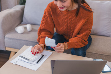 Asian Businesswoman in Orange Sweater Reviews Bills and Calculates Expenses While Using Laptop in...