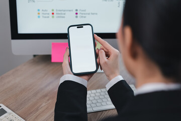 Asian Businesswoman Holding Smartphone With Blank Screen While Working on Computer at Desk.
