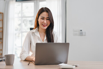 Asian Businesswoman in White Shirt Working on Laptop at Wooden Desk in Bright, Modern Home Office.