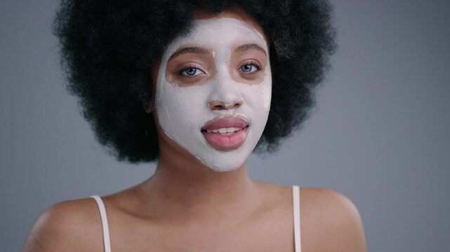 Appealing black young woman enjoying a skin care treatment using a purifying mask for clear face. Portrait of lovely african model with curly hair smiling on grey background.