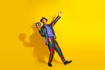 Young man in vibrant colorful suit holding a disco ball against a yellow background, expressing joy...