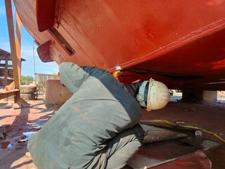A shipyard worker performs metal cutting with a gas welding torch on the underside of a ship's...