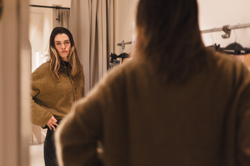 Modern young woman has opportunity to evaluate how she is wearing in fitting room of clothing store. Fashionable clothes. Shopping and pleasure. Girl wear jumper and jeans thinking buy or not.
