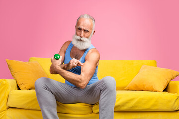 Playful senior man exercising with a dumbbell at home on a sunny yellow couch with a bright pink...