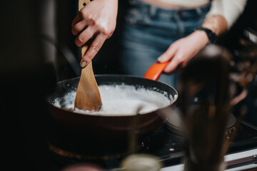 A close-up of hands using a wooden spatula to stir a creamy mixture on a pan. Captures the activity of cooking in a home setting with a focus on preparation and detail.