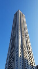 A towering skyscraper stretches towards a clear blue sky, emphasizing its sleek lines and modern design. The sunlight reflects off the glass windows, creating a striking visual impact in the city