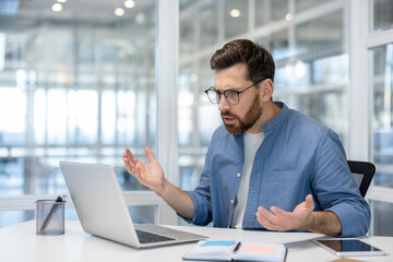 A businessman in a modern office expresses frustration while looking at his laptop, likely...