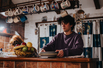 A cozy home kitchen where friends gather. A young man handles plates while another relaxes nearby,...