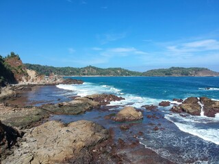 Fototapeta premium Aerial drone view of a rocky coastline with blue ocean waves crashing against the shore under a clear sky
