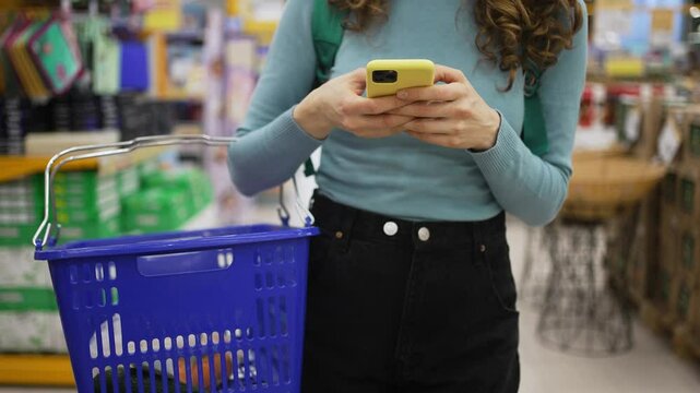 Woman navigating supermarket while checking shopping list on smartphone, holding basket and browsing product displays with digital device