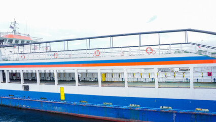 Large passenger and vehicle ferry with white, blue, and red colors sailing on calm ocean waters near the shore, featuring multiple decks, safety lifebuoys, and an open car loading area