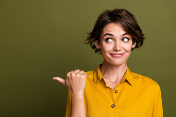 Smiling young woman in yellow shirt pointing with her thumb against a green background, symbolizing...