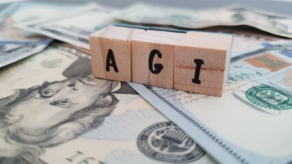 AGI acronym made of wooden letter cubes on pile of US dollar banknotes. Adjusted Gross Income