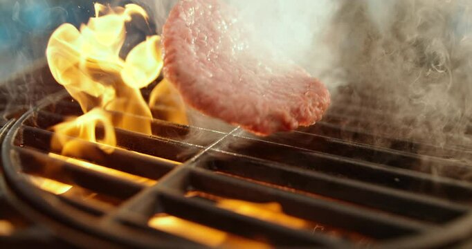 Slow-motion shot of raw beef burger patty dropping on flaming grill to prepare hamburger in the restaurant