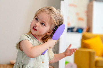 A little blond Caucasian child girl combs her hair with a comb