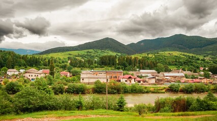 Naklejka premium Picturesque village nestled in green hills by a river under a cloudy sky offers a tranquil landscape.