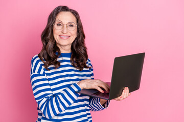 Smiling mature woman in striped sweater holding laptop in front of pink background for advertising...