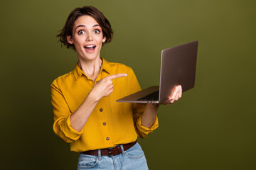 Excited young woman holding and pointing at a laptop while wearing a yellow shirt against a green...
