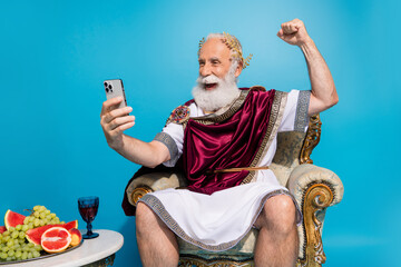 Portrait of delighted rich ancient senator sit throne use phone raise fist isolated on blue color background