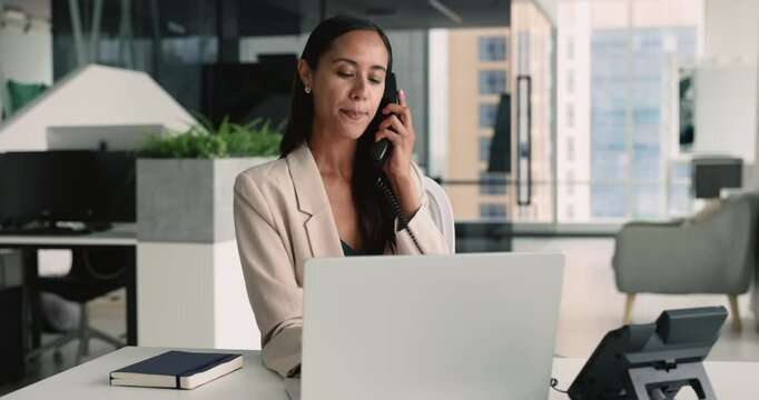 Confident positive busy young professional manager woman working at laptop in successful company office picking up landline phone, talking on telephone, using technology for job communication