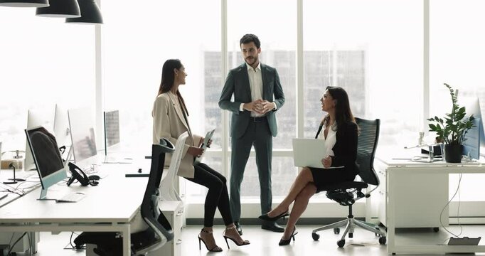 Small diverse business team of three meeting in contemporary urban open office interior, sitting in circle at shared table and large window, talking, discussing teamwork, cooperation