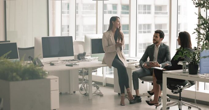Diverse small business team talking in open office space, sitting in circle at shared large workplace table with modern workstations, discussing project, meeting for teamwork, brainstorming