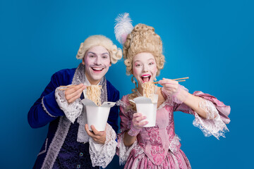 A whimsical couple dressed in elaborate historical costumes enjoys noodle boxes against a vibrant...