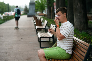 Fototapeta premium Man sitting on a park bench, wiping sweat from his forehead with a tissue while holding sunglasses, feeling the discomfort of summer heat on a sunny day