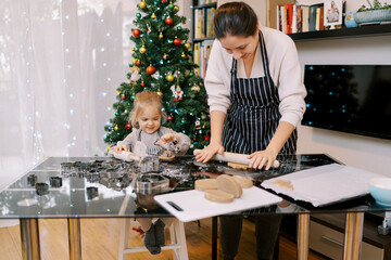 Smiling little girl sitting at the table next to her mother rolling out gingerbread dough