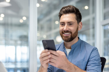 Joyful man uses phone inside office at workplace. Company employee types messages on smartphone and...