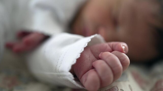 Newborn's tiny hand curled into a fist, dressed in white, resting on a soft surface, with a blurred background conveying the serene and tranquil atmosphere of infancy