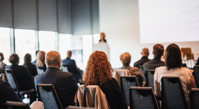 Business and entrepreneurship symposium. Female speaker giving a talk at business meeting. Audience in conference hall. Rear view of unrecognized participant in audience.