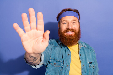 Naklejka premium Portrait of a cheerful bearded man wearing a denim jacket and headband against a vibrant purple background, giving a happy wave