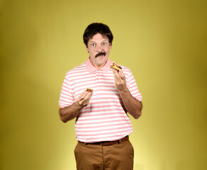 Young adult with moustache, polo shirt and trousers, looking at camera and eating a cake with satisfied face.