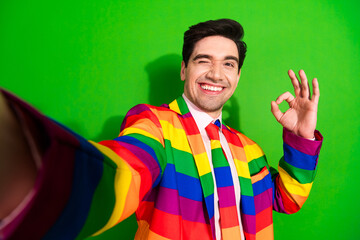 Young man in a vibrant rainbow suit winking and gesturing OK against a bright green background,...