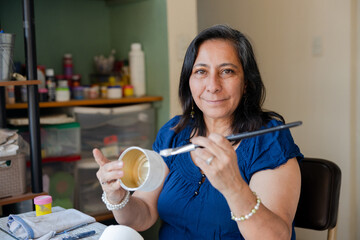 Portrait of hispanic woman doing crafts at home - senior woman smiling at the camera while painting...