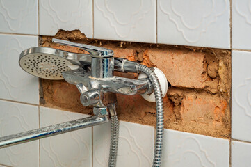 A corroded chrome shower faucet is mounted on a damaged tiled wall, exposing brick and mortar.