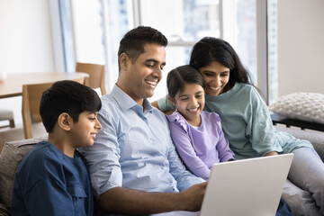 Indian family with children sits together on cozy sofa with laptop, watching movie, entertaining or educational content with interest and engagement, enjoying joint hobby, electronic shopping at home