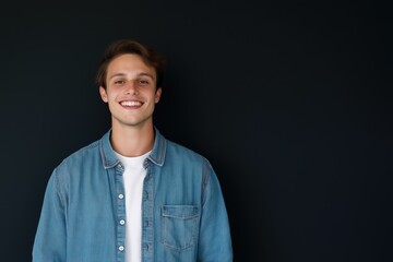 Obraz premium A young man is smiling and wearing a blue denim jacket. He is standing in front of a black background